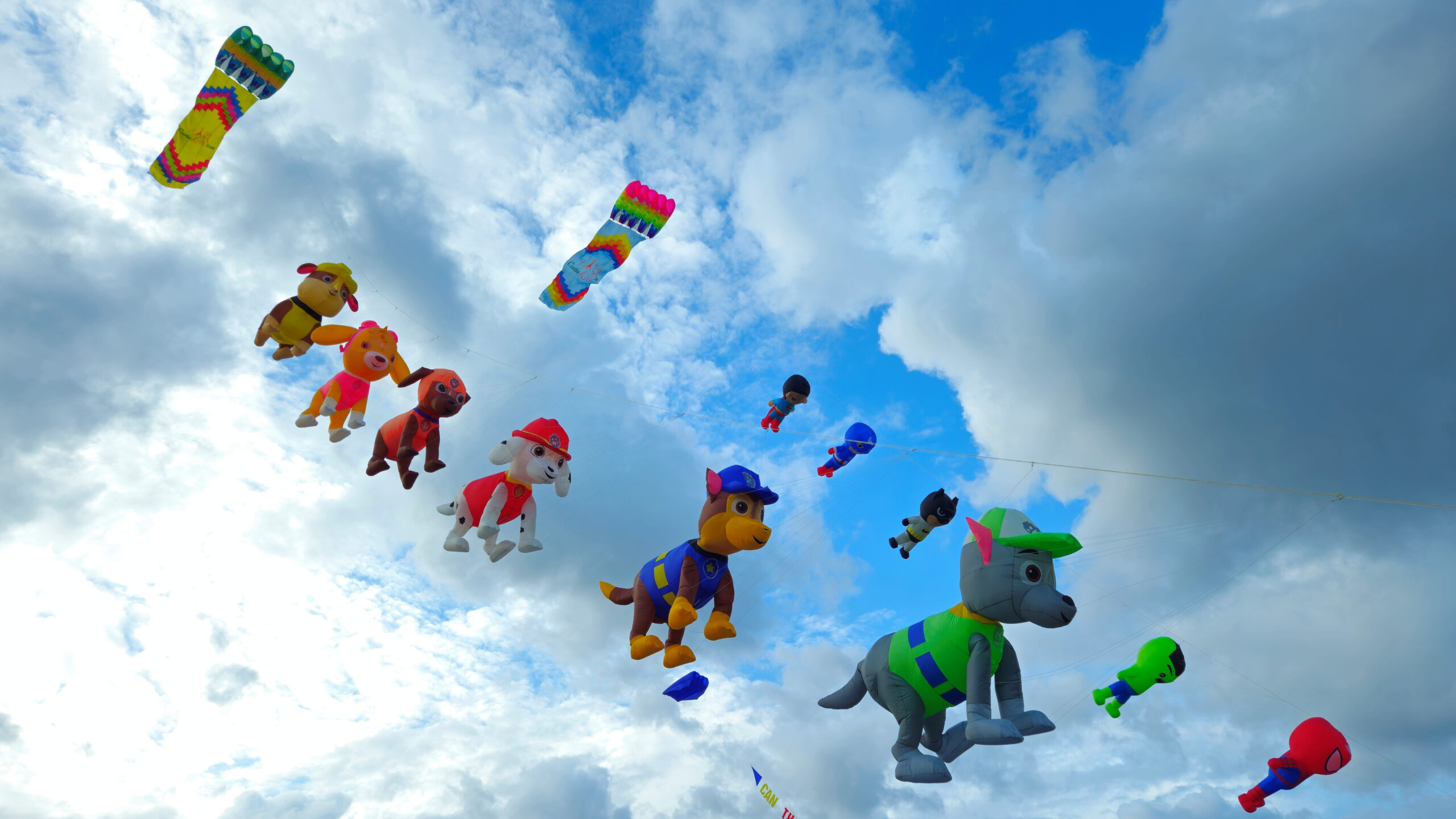A group of colorful kites, shaped like cartoon characters and rectangular, is flying in the bright blue sky.